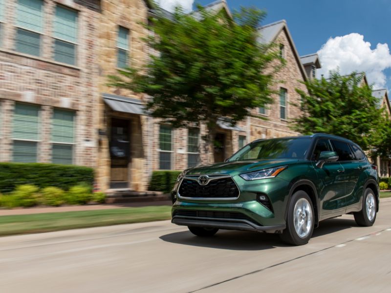 2023 metallic green Toyota Highlander driving in a densely populated suburban area